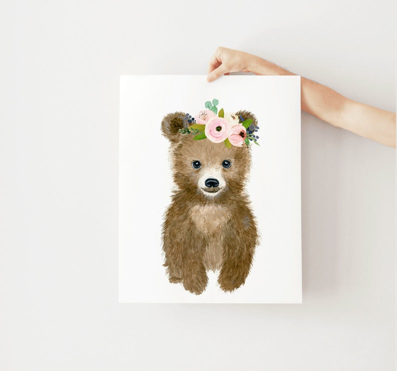 May include: A brown bear cub with a pink and green floral crown. The bear is looking at the camera with a sweet expression.