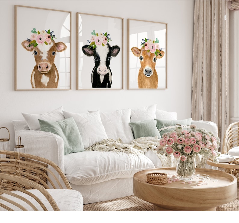 Puede incluir: Tres pinturas de acuarela enmarcadas de vacas con coronas de flores. Las vacas son marrones, negras y blancas, y marrones. Las pinturas est&aacute;n colgadas en una pared blanca sobre un sof&aacute; blanco.