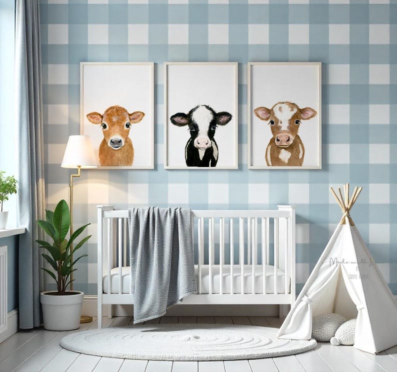May include: Three watercolor paintings of cow faces in white frames against a blue and white gingham wallpaper. The paintings are hanging above a white crib with a gray blanket and a white teepee tent.