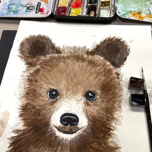 May include: Watercolor painting of a brown bear cub with big, dark eyes. The bear is looking directly at the viewer with a sweet expression. The painting is on white paper and there are watercolor paints and a brush in the background.