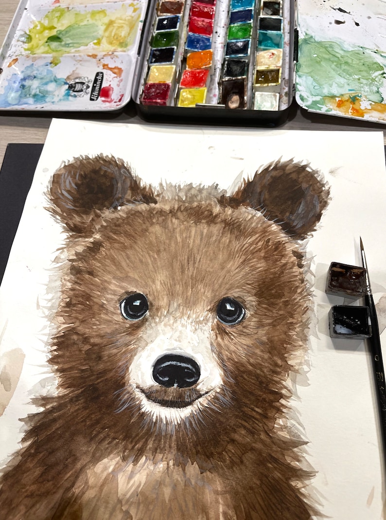 May include: Watercolor painting of a brown bear cub with big, dark eyes. The bear is looking directly at the viewer with a sweet expression. The painting is on white paper and there are watercolor paints and a brush in the background.