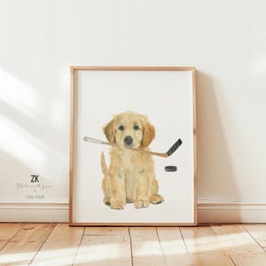 May include: Watercolor print of a golden retriever puppy with a hockey stick in its mouth and a black hockey puck. The artwork is framed in a light brown wooden frame and set against a white background.