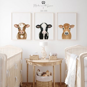May include: Three framed watercolor paintings of brown and white calves with different colored markings. The paintings are hung on a white wall above a wooden table with a lamp and two wicker baskets. The table is between two white cribs with white bedding.