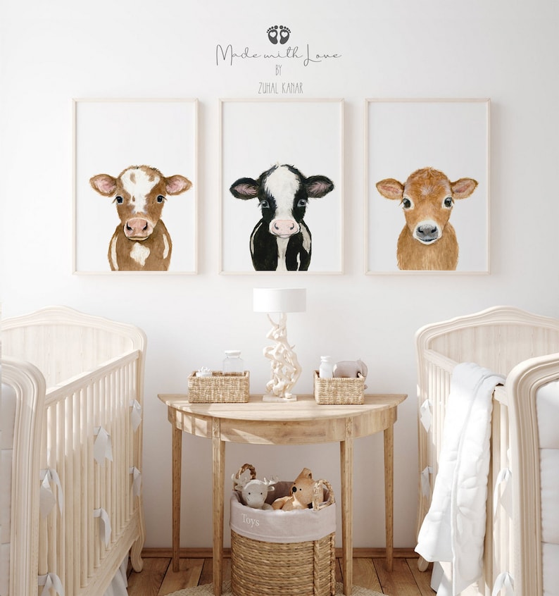 May include: Three framed watercolor paintings of brown and white calves with different colored markings. The paintings are hung on a white wall above a wooden table with a lamp and two wicker baskets. The table is between two white cribs with white bedding.