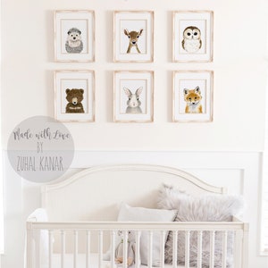 May include: A white crib with a white mattress and two white pillows. The crib is decorated with six framed prints of woodland animals, including a hedgehog, deer, owl, bear, rabbit, and fox. The prints are arranged in two rows of three. The text "Born to be Wild" is written above the prints. The text "Made with Love by Zuhal Kanar" is written below the prints.