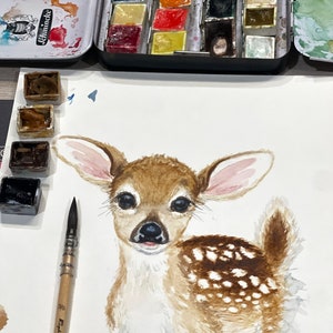 May include: Watercolor painting of a fawn with brown and white spots. The fawn has large ears and big brown eyes. The painting is on white paper and there are watercolor paints and a paintbrush in the background.