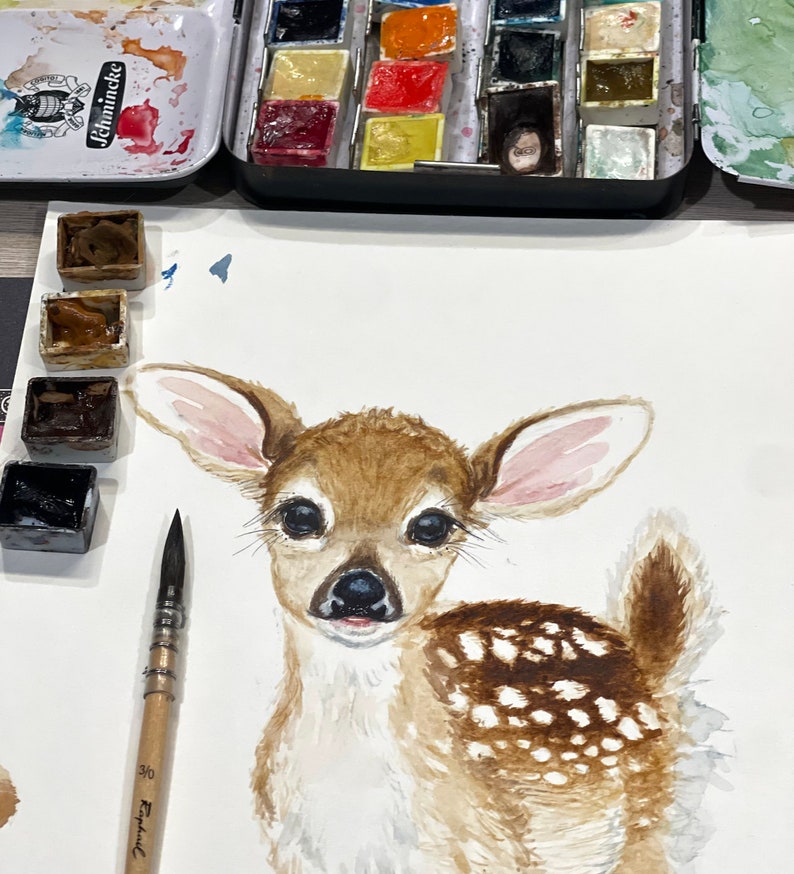 May include: Watercolor painting of a fawn with brown and white spots. The fawn has large ears and big brown eyes. The painting is on white paper and there are watercolor paints and a paintbrush in the background.
