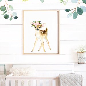 May include: A watercolor painting of a fawn with a pink flower crown on a white background. The fawn is standing on all four legs and looking to the right. The painting is framed in a light wood frame.