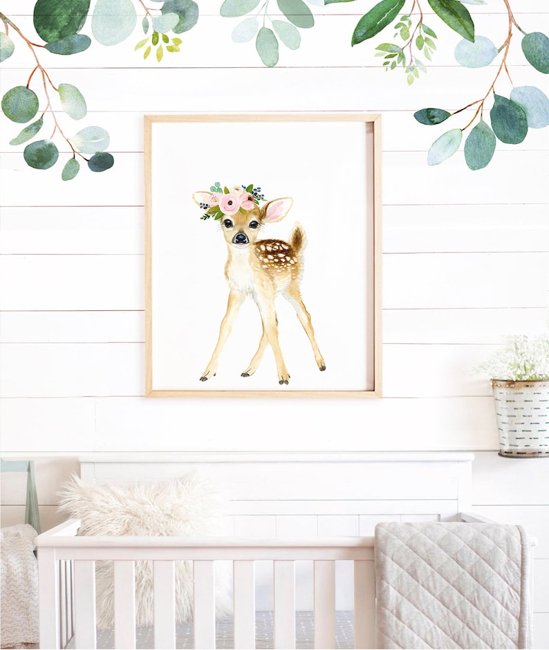 May include: A watercolor painting of a fawn with a pink flower crown on a white background. The fawn is standing on all four legs and looking to the right. The painting is framed in a light wood frame.
