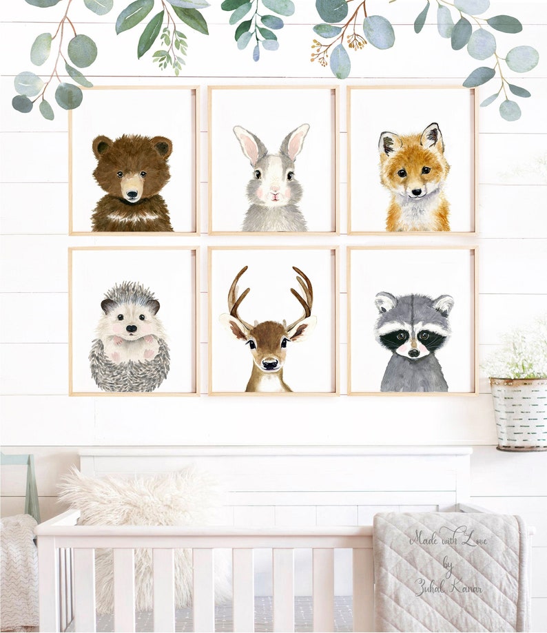 May include: Six framed watercolor paintings of woodland animals, including a bear, rabbit, fox, hedgehog, deer, and raccoon. The paintings are displayed on a white wall with a crib and a blanket in the foreground.