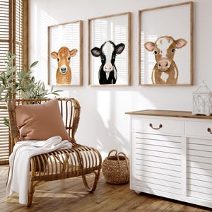 May include: Three framed watercolor paintings of cow faces. The paintings are hung on a white wall above a white dresser with a wooden top. A wicker chair with a brown throw blanket and a brown pillow sits in front of the dresser.