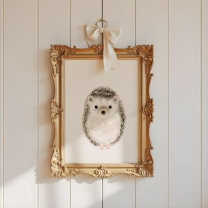 Op de afbeelding: Een ingelijste print van een aquarel illustratie van een egel. De egel is grijs en wit met zwarte ogen en een roze neus. De lijst is goudkleurig met sierlijke details.