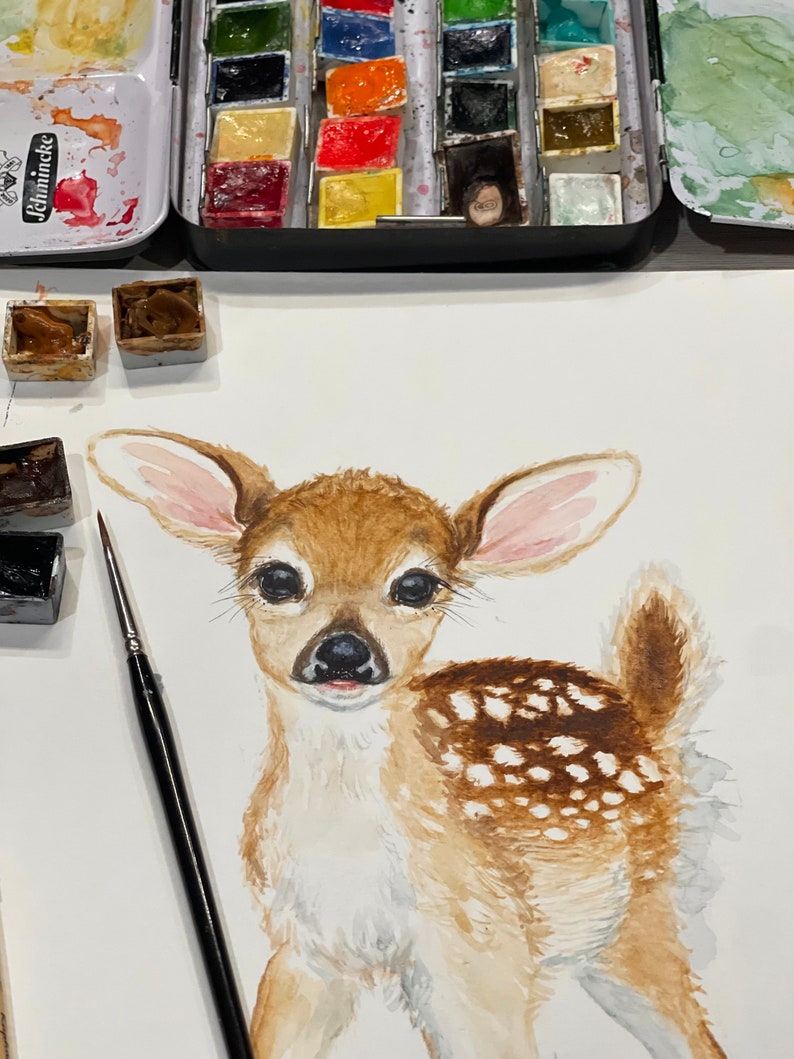 May include: Watercolor painting of a fawn with brown and white fur. The fawn has large, dark eyes and a pink nose. The painting is on a white background.
