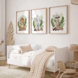 May include: Three framed watercolor prints featuring a lion, elephant, and meerkat surrounded by green foliage. The prints are displayed on a white wall above a white daybed with a beige blanket and pillows.