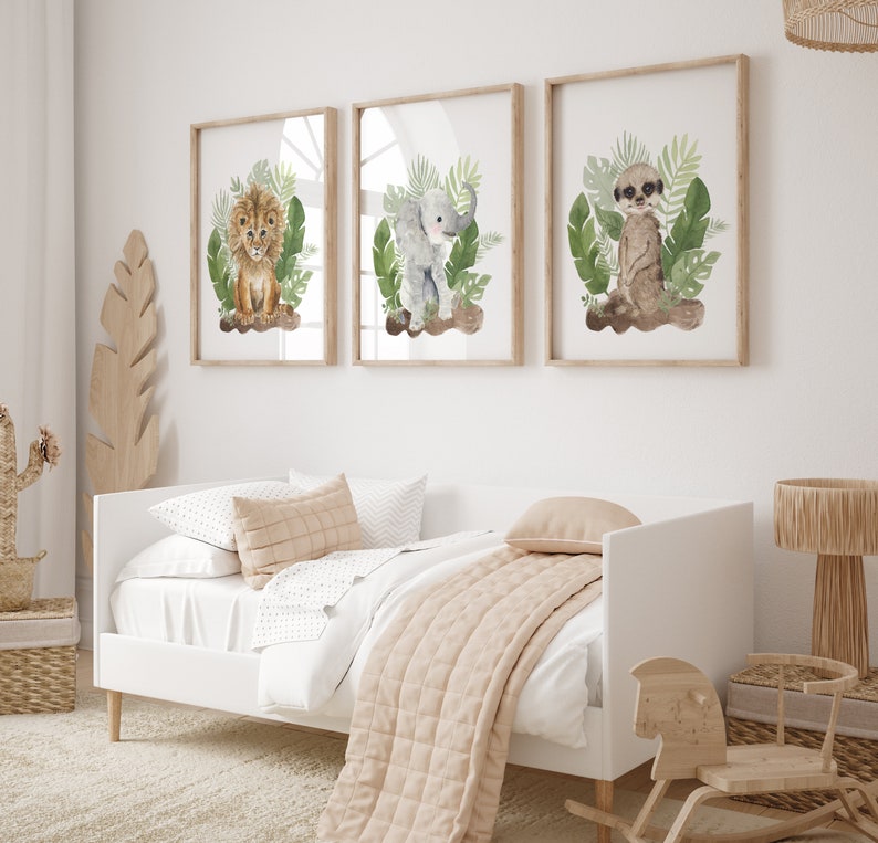 May include: Three framed watercolor prints featuring a lion, elephant, and meerkat surrounded by green foliage. The prints are displayed on a white wall above a white daybed with a beige blanket and pillows.