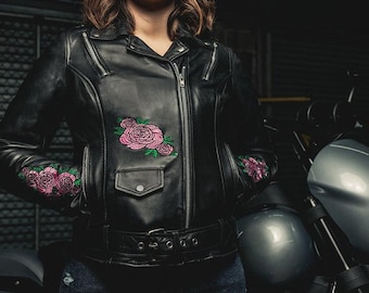 May include: Black leather jacket with pink rose embroidery on the front and sleeves. The jacket features a classic biker style with a zippered front, pockets, and a belted waist. The jacket is shown with a motorcycle in the background.