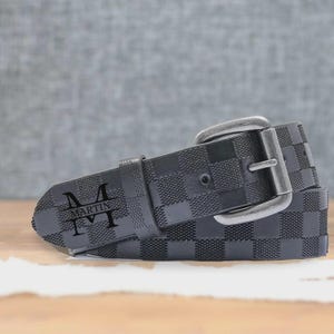 May include: Black checkered belt with a silver buckle. The belt features the name "MARTIN" with the letter "M" on each side of the name. The belt is made of a textured material and is displayed on a wooden surface.
