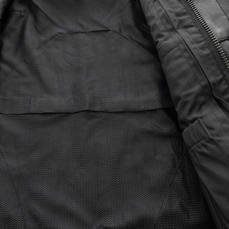 May include: Close-up of the interior of a black jacket, revealing a textured, mesh-like lining. The jacket's interior features a pocket and a zipper detail, showcasing the garment's construction and design.