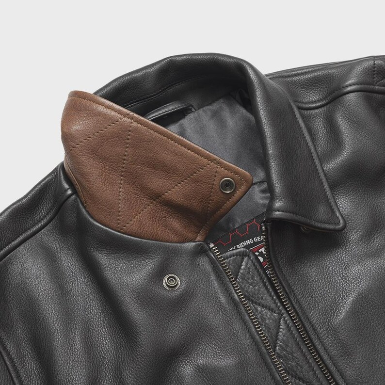 May include: Close-up of a black leather jacket with a brown leather collar. The collar has a diamond-stitched pattern. The jacket features a zipper closure and snap details. The overall design is classic and rugged.