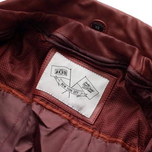 Puede incluir: Primer plano del interior de una chaqueta de cuero burdeos. Una etiqueta blanca con el texto "FIRST CO." y "NEW YORK CITY" es visible. La chaqueta tiene un forro de malla y una etiqueta de talla "XS".