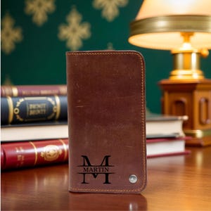 May include: A brown leather wallet with the name "MARTIN" and the initial "M" engraved on the front. The wallet is rectangular and appears to be closed. The background includes books and a lamp.