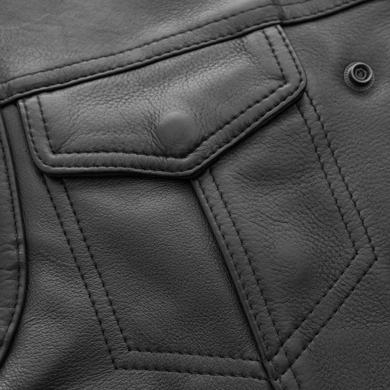May include: Close-up of a black leather jacket detail. The image shows a pocket with a button closure, decorative stitching, and a snap closure. The leather has a textured appearance.