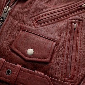 Puede incluir: Primer plano de una chaqueta de cuero burdeos. Incluye un bolsillo con cremallera, un bolsillo con cierre de bot&oacute;n a presi&oacute;n y un cintur&oacute;n con hebilla. La chaqueta tiene una superficie texturizada y detalles de costura visibles.
