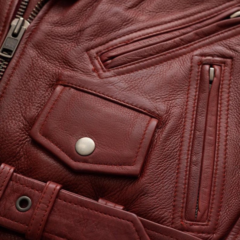 Puede incluir: Primer plano de una chaqueta de cuero burdeos. Incluye un bolsillo con cremallera, un bolsillo con cierre de bot&oacute;n a presi&oacute;n y un cintur&oacute;n con hebilla. La chaqueta tiene una superficie texturizada y detalles de costura visibles.