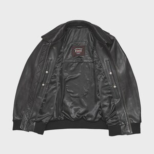 May include: Black leather jacket with a classic bomber style. The jacket features a collar, snap closures, and a ribbed hem and cuffs. The interior lining is black, and a small label reads "FIRST".