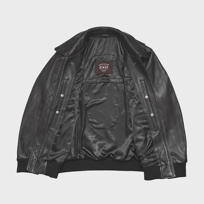 May include: Black leather jacket with a classic bomber style. The jacket features a collar, snap closures, and a ribbed hem and cuffs. The interior lining is black, and a small label reads "FIRST".