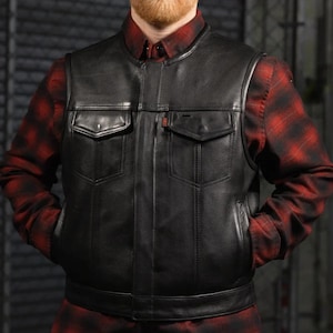 May include: Black leather vest with two chest pockets and two side pockets. The vest has a round collar and a button closure. The person is wearing a red and black plaid shirt underneath the vest.