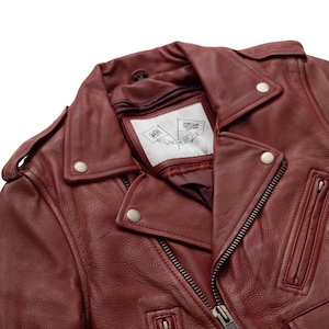 Puede incluir: Primer plano de una chaqueta de cuero burdeos con un estilo motero cl&aacute;sico. La chaqueta presenta un cuello con muescas, herrajes plateados y una cremallera frontal. La etiqueta interior muestra un gr&aacute;fico de bandera y el texto "NEW YORK CITY".