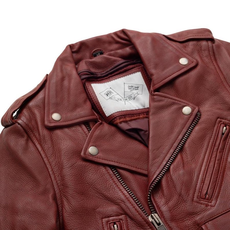 Puede incluir: Primer plano de una chaqueta de cuero burdeos con un estilo motero cl&aacute;sico. La chaqueta presenta un cuello con muescas, herrajes plateados y una cremallera frontal. La etiqueta interior muestra un gr&aacute;fico de bandera y el texto "NEW YORK CITY".