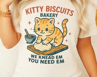 Könnte beinhalten: Cremefarbenes T-Shirt mit einem orangefarbenen getigerten Kätzchen, das Teig auf einem Holzbrett knetet. Der Text lautet "KITTY BISCUITS BAKERY" und "WE KNEAD EM YOU NEED EM". Das Design enthält Sterne und eine Schüssel.