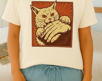 May include: Cream-colored t-shirt featuring a cartoon cat illustration in a brown square. The cat is light yellow with red eyes and is biting a hand. The person is wearing blue shorts.