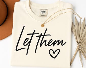 May include: A cream colored t-shirt with the text "Let them" in black script with a small heart at the end. The shirt is folded and laid flat on a white surface.