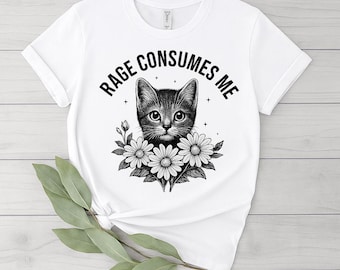 Op de afbeelding: Wit T-shirt met een zwart-wit illustratie van een katje omringd door madeliefjes. De tekst "RAGE CONSUMES ME" staat boven het hoofd van de kat. Het shirt heeft korte mouwen en een ronde hals.