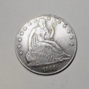 May include: A silver coin featuring a seated figure holding a staff and shield, surrounded by stars. The coin is dated 1864 and has a detailed design. The coin is on a gray background.