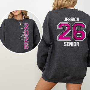 Pu&ograve; includere: Una felpa grigio scuro con il nome "Jessica" e il numero "26" in rosa acceso, bordato di bianco. La parola "Senior" &egrave; stampata sotto. La parte anteriore della felpa ha il testo "Class of 2026".