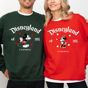 Pode incluir: Duas camisolas, uma verde e outra vermelha, com o log&oacute;tipo da Disneyland com Mickey e Minnie Mouse. A camisola verde tem o Mickey Mouse, e a camisola vermelha tem a Minnie Mouse. Ambas t&ecirc;m o texto "est 1955 CALIFORNIA".
