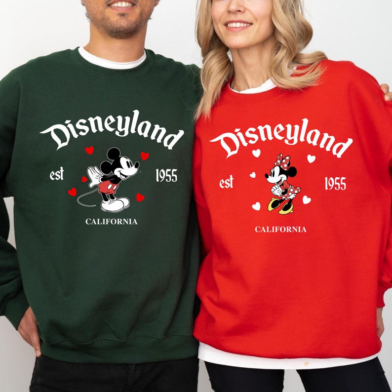 Pode incluir: Duas camisolas, uma verde e outra vermelha, com o log&oacute;tipo da Disneyland com Mickey e Minnie Mouse. A camisola verde tem o Mickey Mouse, e a camisola vermelha tem a Minnie Mouse. Ambas t&ecirc;m o texto "est 1955 CALIFORNIA".