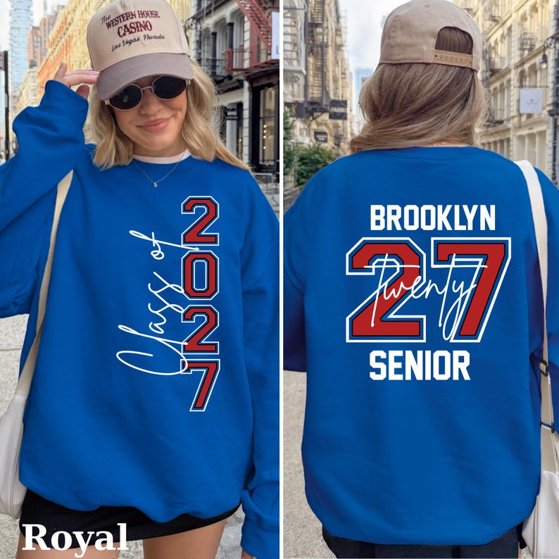 May include: Royal blue sweatshirt with white and red lettering. The front reads "Class of 2027" vertically. The back says "Brooklyn 27 Twenty Senior". The sweatshirt is a classic crew neck style.