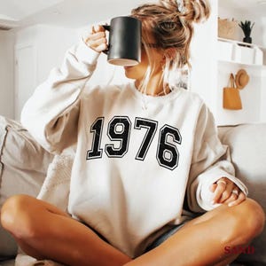 Könnte beinhalten: Cremefarbenes Sweatshirt mit der Jahreszahl "1976" in großen, schwarzen Blockbuchstaben. Das Sweatshirt hat lange Ärmel und einen Rundhalsausschnitt. Eine schwarze Tasse wird vor dem Gesicht gehalten.