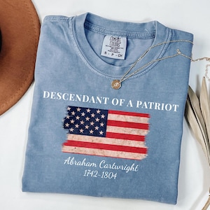 May include: A light blue t-shirt with the text "DESCENDANT OF A PATRIOT" above a stylized American flag. Below the flag is the name "Abraham Cartwright" and the years "1742-1804". A gold necklace with a pendant rests on the shirt.