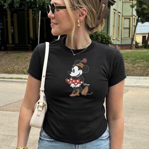 Może przedstawiać: Czarny t-shirt z grafiką Myszki Minnie. Postać z kresk&oacute;wki ma na sobie czerwoną sukienkę w kropki i kapelusz z kwiatkiem. Koszulka ma kr&oacute;tkie rękawy i jest połączona z niebieskimi dżinsami.