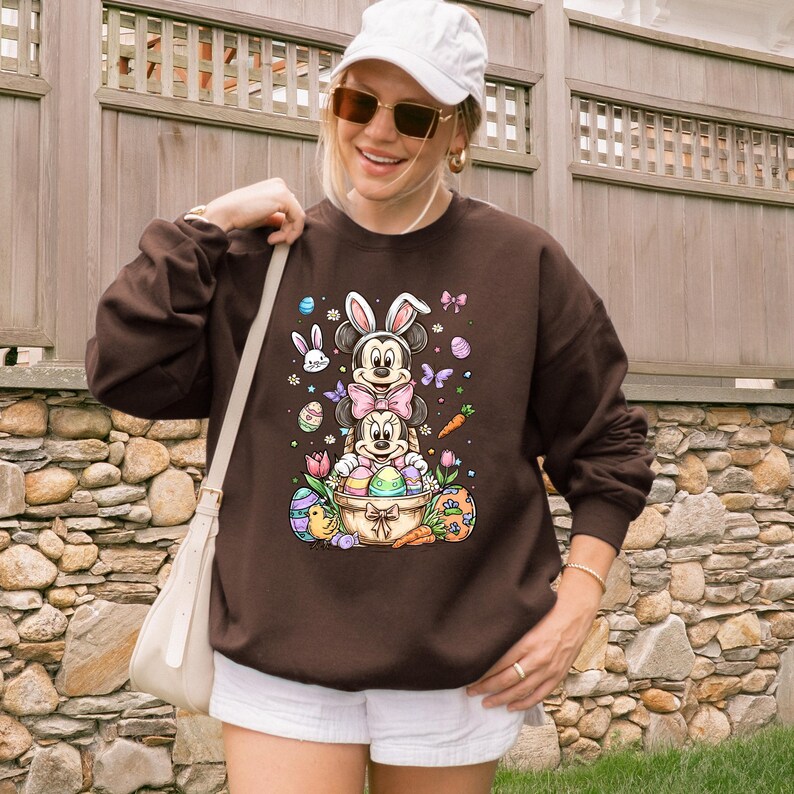 K&ouml;nnte beinhalten: Ein braunes Sweatshirt mit einem farbenfrohen Ostermotiv. Die Grafik zeigt Mickey- und Minnie-Mouse-Figuren mit Hasenohren, Ostereiern, Schmetterlingen und Karotten. Das Sweatshirt hat eine einfarbige braune Farbe.