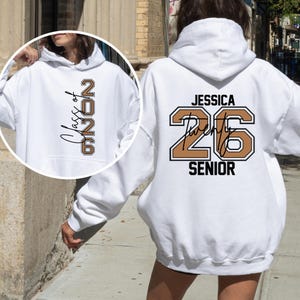 Pu&ograve; includere: Felpa con cappuccio bianca con tasca frontale e cappuccio. La parte anteriore presenta il testo "Class of 2026" in un design verticale. Il retro mostra "Jessica 26 Senior" in marrone e nero.