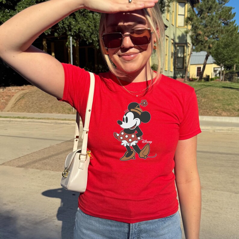 Może przedstawiać: Czerwony t-shirt z grafiką Myszki Minnie. Postać z kresk&oacute;wki jest przedstawiona w klasycznej pozie, w sp&oacute;dnicy w kropki i kapeluszu z kwiatkiem. Koszulka jest połączona z niebieskimi dżinsami i białą torebką.
