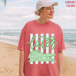May include: A red-orange t-shirt with a light green alligator design and the word "later" in a cursive font. The shirt has vertical light blue stripes behind the text and alligator. The words "COMFORT COLORS" are in the upper right corner.