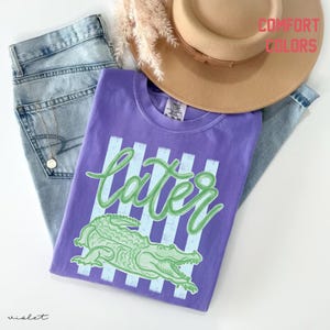 May include: A purple t-shirt with the word "later" in green script and a green alligator graphic. The shirt is paired with blue jeans, a tan hat, and a necklace. The words "Comfort Colors" are printed in pink.